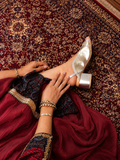 A close-up lifestyle shot of a woman wearing a metallic silver pointed-toe flat shoe with a block heel, styled with a deep red traditional dress and gold jewelry, sitting on a patterned red rug.